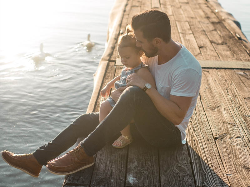 Father embraces young toddler while sitting on the edge of a wharf